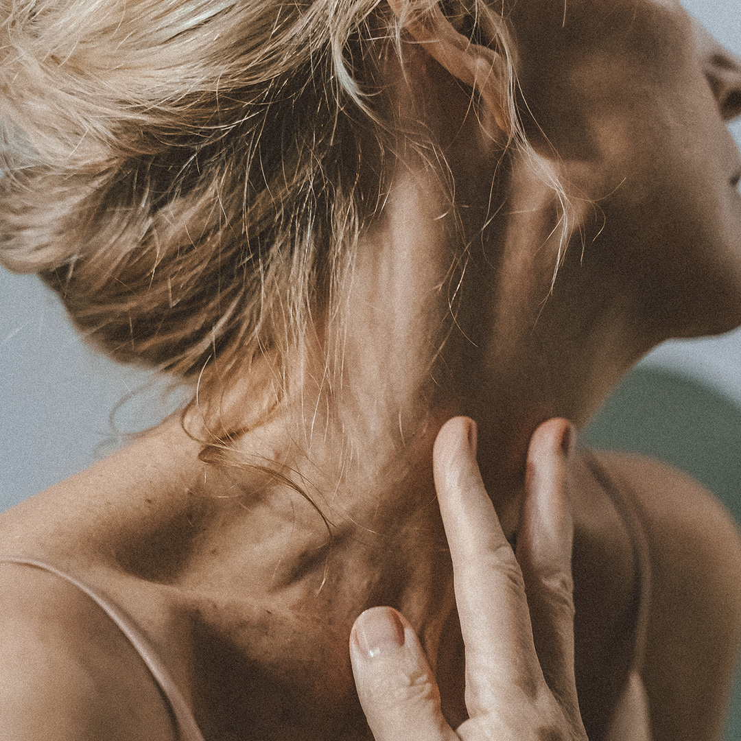 Nahaufnahme einer Person, die sanft mit der Hand ihren Hals berührt. Die Person hat helles Haar, das zu einem lockeren Knoten hochgebunden ist. Ihre Haut wirkt weich und sie trägt ein ärmelloses Oberteil, was einen intimen und ruhigen Moment einfängt.