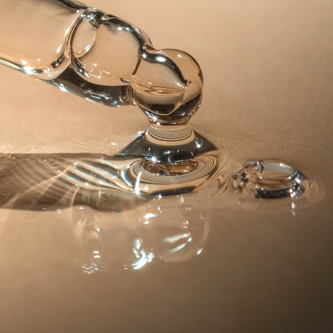 Nahaufnahme einer Glaspipette, die einen Flüssigkeitstropfen auf eine glatte Oberfläche abgibt. Sowohl die klare Flüssigkeit als auch die Pipette reflektieren das Licht und erzeugen komplizierte Muster und Reflexionen. Der Hintergrund ist sanft beleuchtet, wodurch die Transparenz der Pipette und der Flüssigkeit hervorgehoben wird.