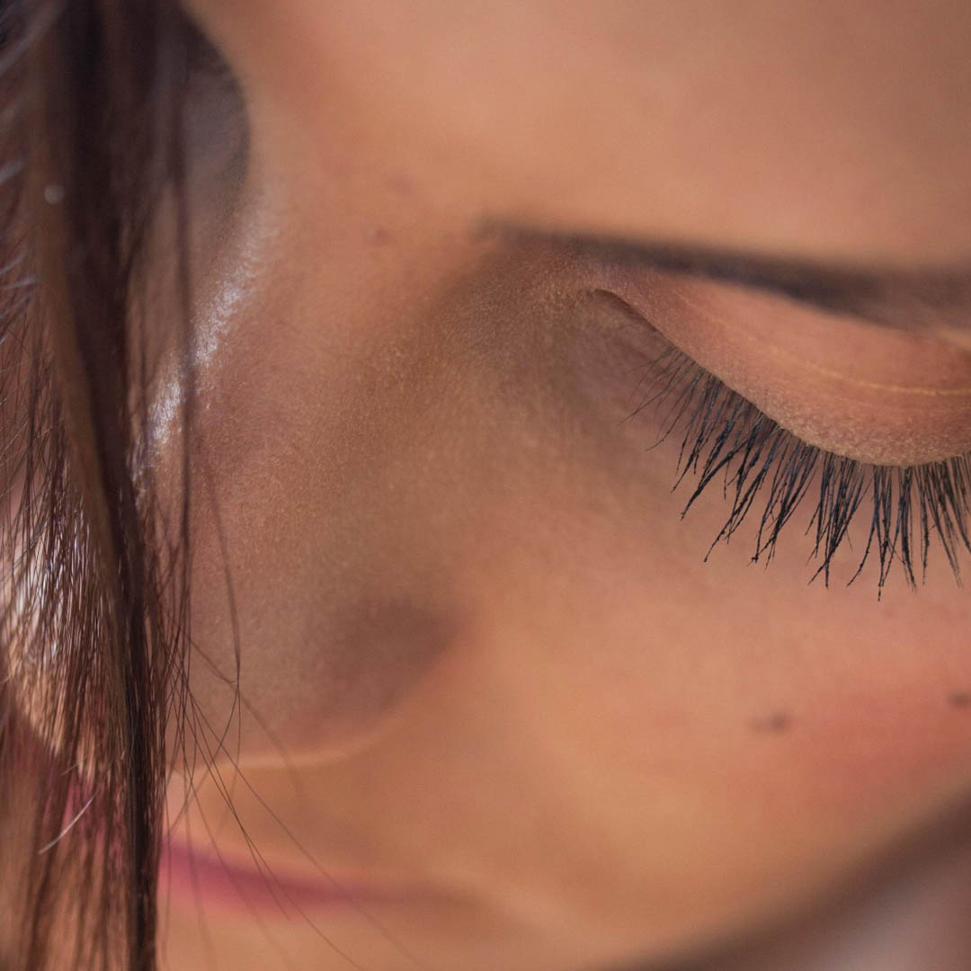 Nahaufnahme des Gesichts einer Person mit Fokus auf die Augenpartie. Die Wimpern sind lang und dunkel, ein Auge ist teilweise geschlossen. Weiches Licht hebt die Haut und die feinen Gesichtszüge hervor und schafft eine intime und heitere Atmosphäre. Ein paar Haarsträhnen sind sichtbar.