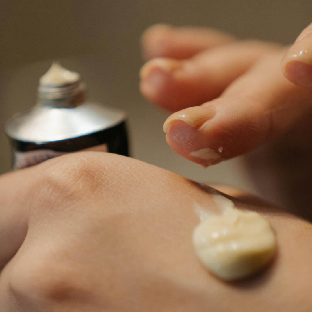 Eine Hand mit einem kleinen Klecks Creme wird aus einer Metalltube gedrückt. Ein anderer Finger berührt die Creme, was darauf schließen lässt, dass das Produkt auf die Haut aufgetragen oder verteilt wird. Der Hintergrund ist unscharf.