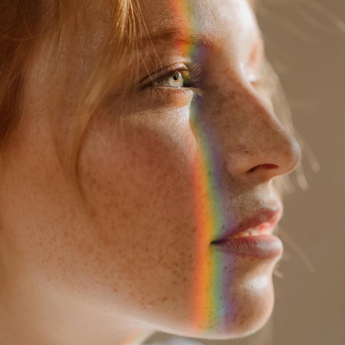 Eine Nahaufnahme des Profils einer Person mit Sommersprossen, teilweise beleuchtet durch einen Regenbogen, der ihr Gesicht überzieht. Der Blick der Person ist nach oben gerichtet und das warme Licht betont die Details ihrer Haut und Haare.