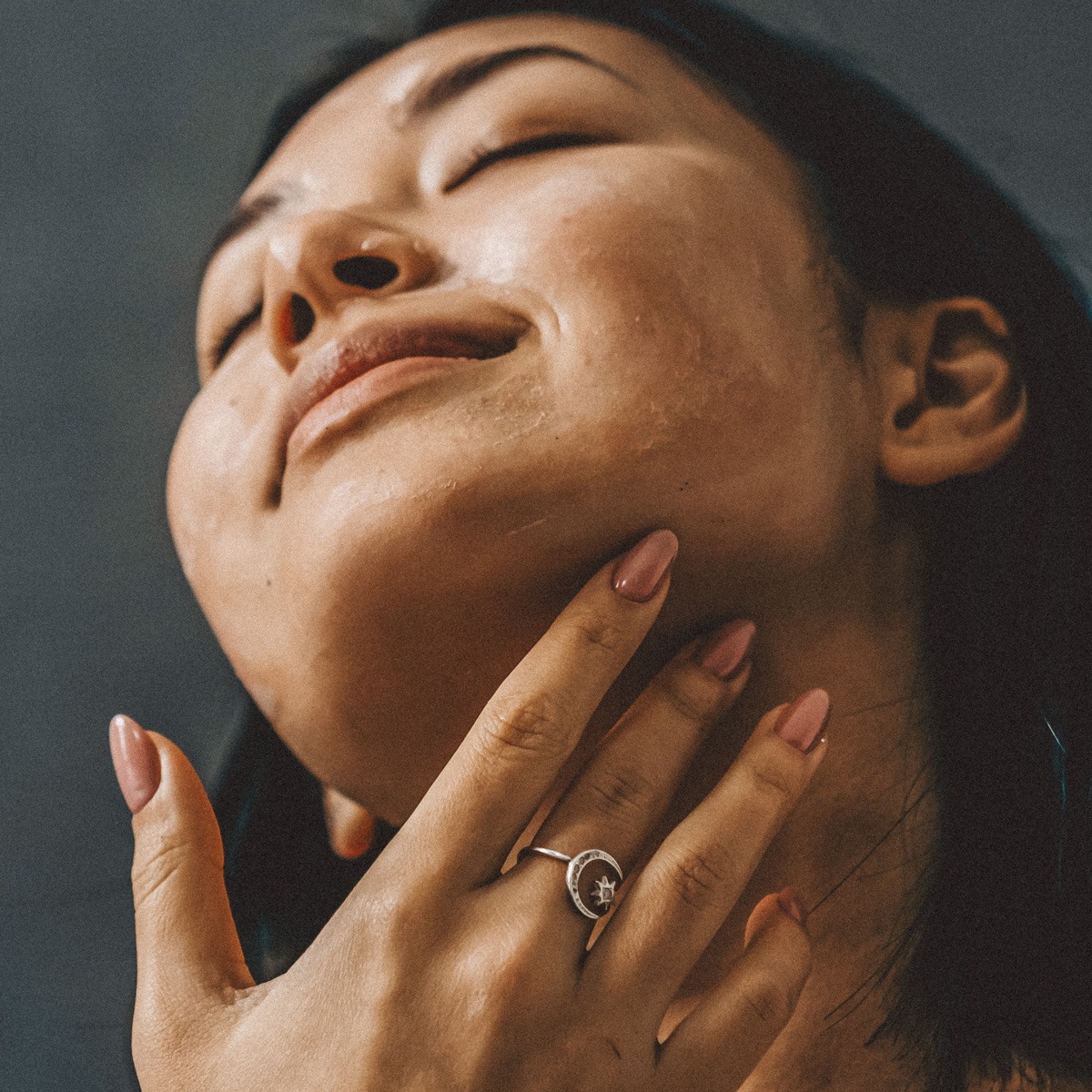 Eine Person mit langem, dunklem Haar schließt die Augen, lächelt sanft und berührt dabei sanft ihren Hals. Sie hat glatte Haut und trägt einen silbernen Ring mit einem kleinen Muster darauf. Ihre Nägel sind manikürt und hellrosa lackiert.
