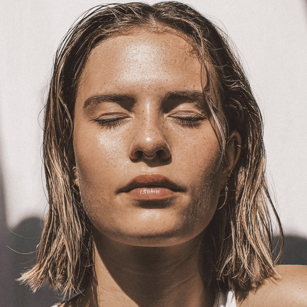 Eine Nahaufnahme einer Person mit kurzen, nassen Haaren und geschlossenen Augen, die mit heiterem Gesichtsausdruck ein Sonnenbad nimmt. Ihr Gesicht wird vom natürlichen Sonnenlicht beleuchtet, wodurch ihre Haut und Gesichtszüge hervorgehoben werden. Der Hintergrund ist verschwommen und zeigt eine helle Wand.