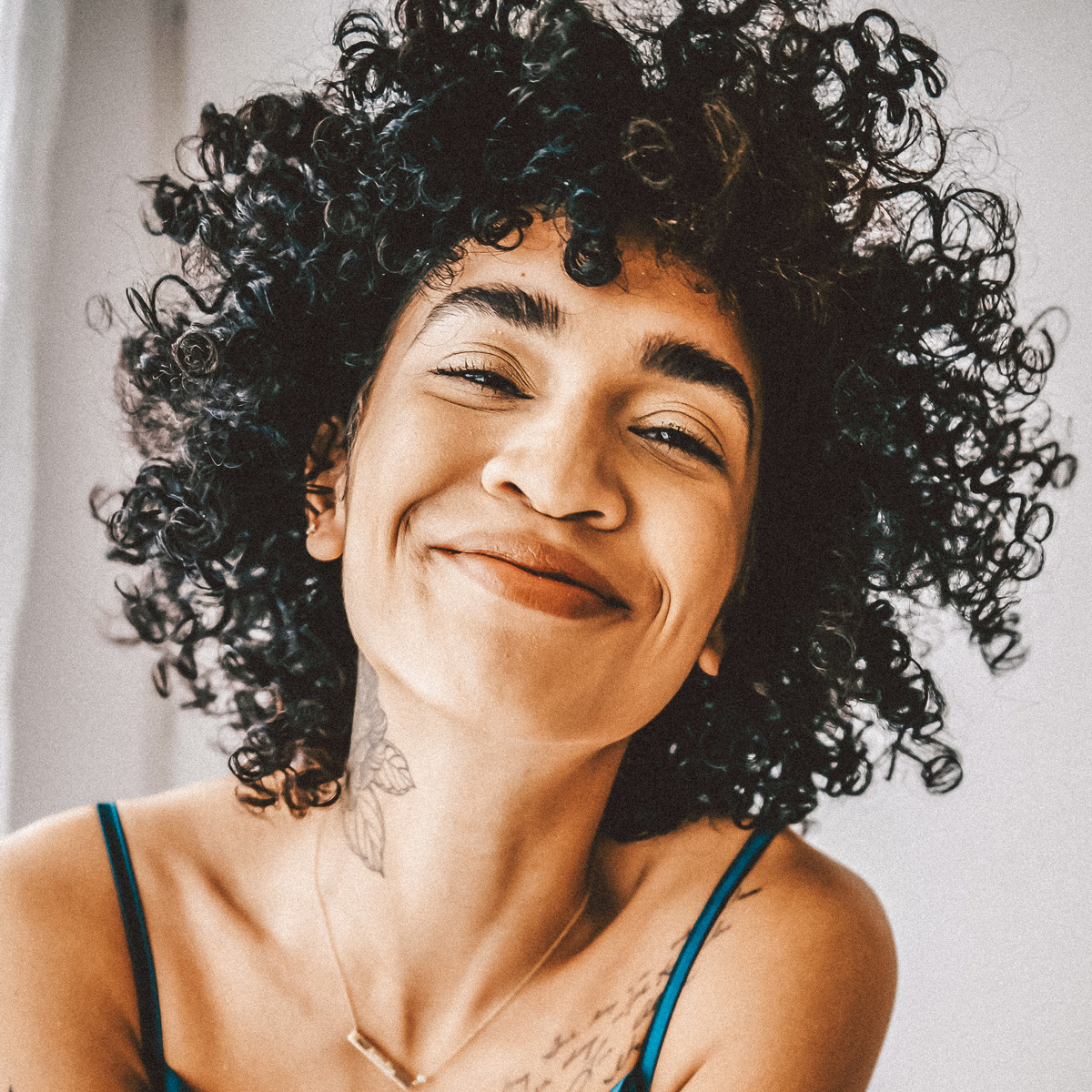 Eine Person mit lockigem Haar lächelt warm in die Kamera. Sie trägt ein ärmelloses Oberteil und zeigt ein Tattoo auf Schulter und Hals. Der Hintergrund ist sanft beleuchtet, was ihren freudigen Ausdruck und die natürliche Textur ihrer Haare betont.