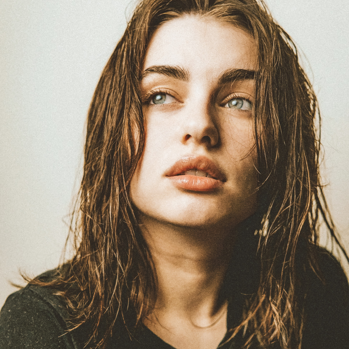 Eine Nahaufnahme einer jungen Frau mit nassem, schulterlangem Haar, die leicht nach oben und zur Seite blickt. Sie hat helle Augen, volle Lippen und trägt ein dunkles Oberteil. Der Hintergrund ist schlicht und in einem neutralen Ton gehalten.