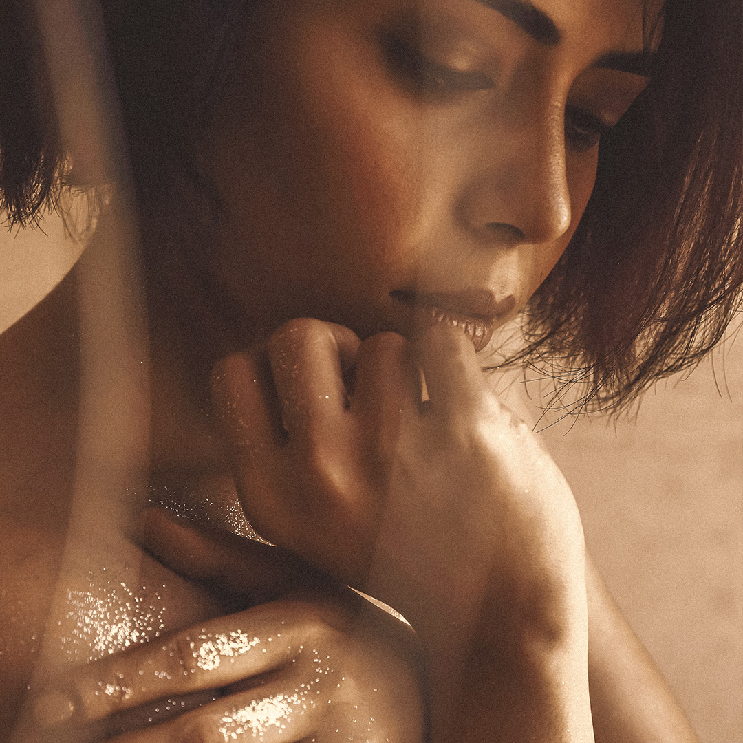 Nahaufnahme einer Frau mit kurzen braunen Haaren, die teilweise mit goldenem Glitzer bedeckt sind. Sie blickt nachdenklich nach unten und berührt mit der Hand sanft ihr Kinn und ihre Schulter. Der Hintergrund ist unscharf, was dem Bild eine weiche, intime Atmosphäre verleiht.